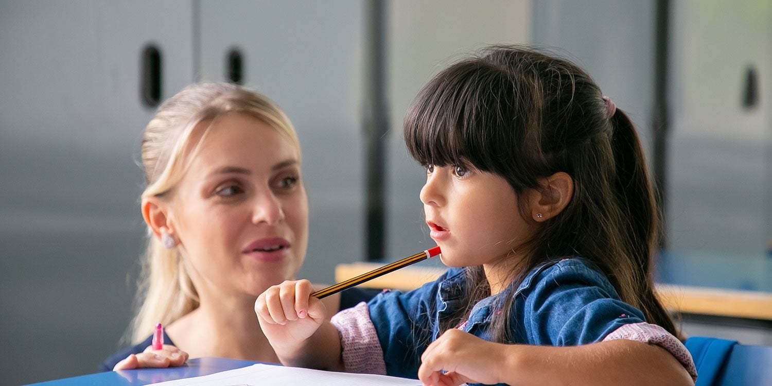 Adult and child at desk, child holding pencil to mouth.