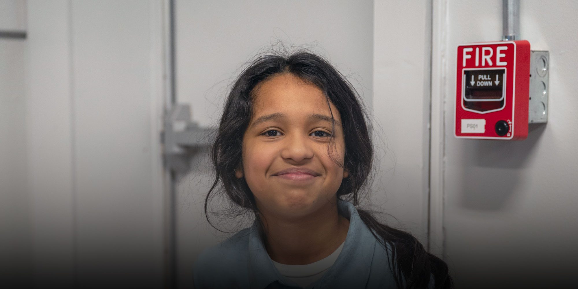 Smiling student with long dark hair in a school hallway.