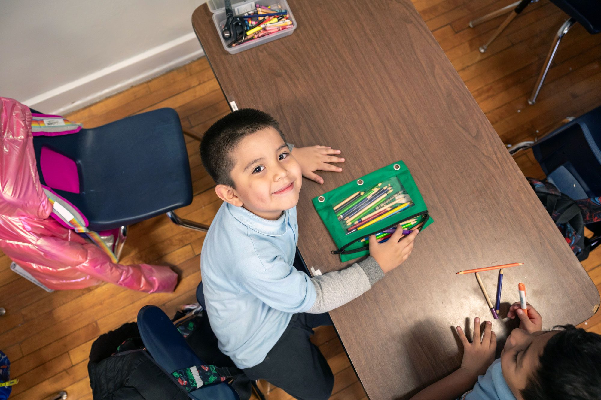 Overhead view of a smiling student at a classroom table with a pouch of colored pencils.